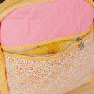 A close-up of a quilted fabric backpack or bag with a pink diamond-quilted top panel, yellow gingham trim, and a floral front pocket on a light wood surface.