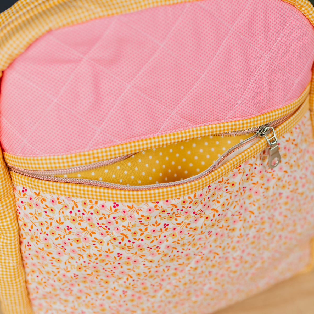A close-up of a quilted fabric backpack or bag with a pink diamond-quilted top panel, yellow gingham trim, and a floral front pocket on a light wood surface.
