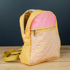A small fabric backpack with a yellow gingham and floral patterned body, a pink interior lining, and a yellow shoulder strap, standing on a light wood surface against a dark gray background.