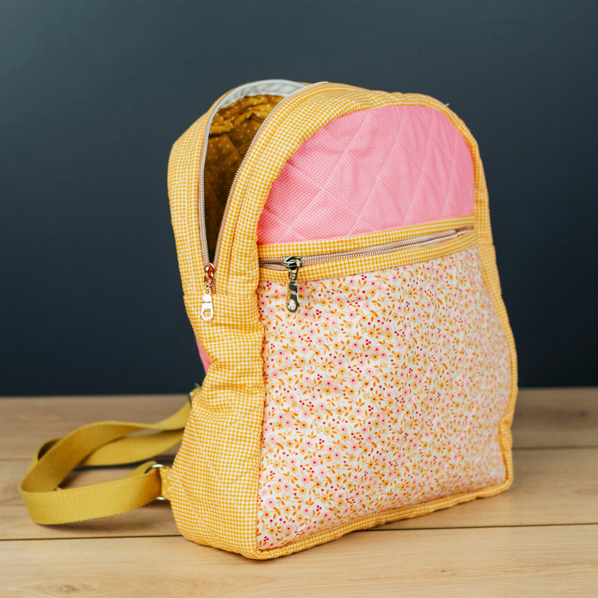 A small fabric backpack with a yellow gingham and floral patterned body, a pink interior lining, and a yellow shoulder strap, standing on a light wood surface against a dark gray background.