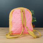 A pink quilted backpack with mustard yellow straps and trim, standing on a light wood surface against a dark gray background.