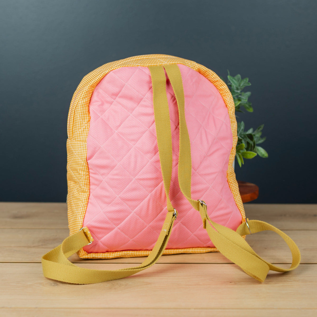A pink quilted backpack with mustard yellow straps and trim, standing on a light wood surface against a dark gray background.