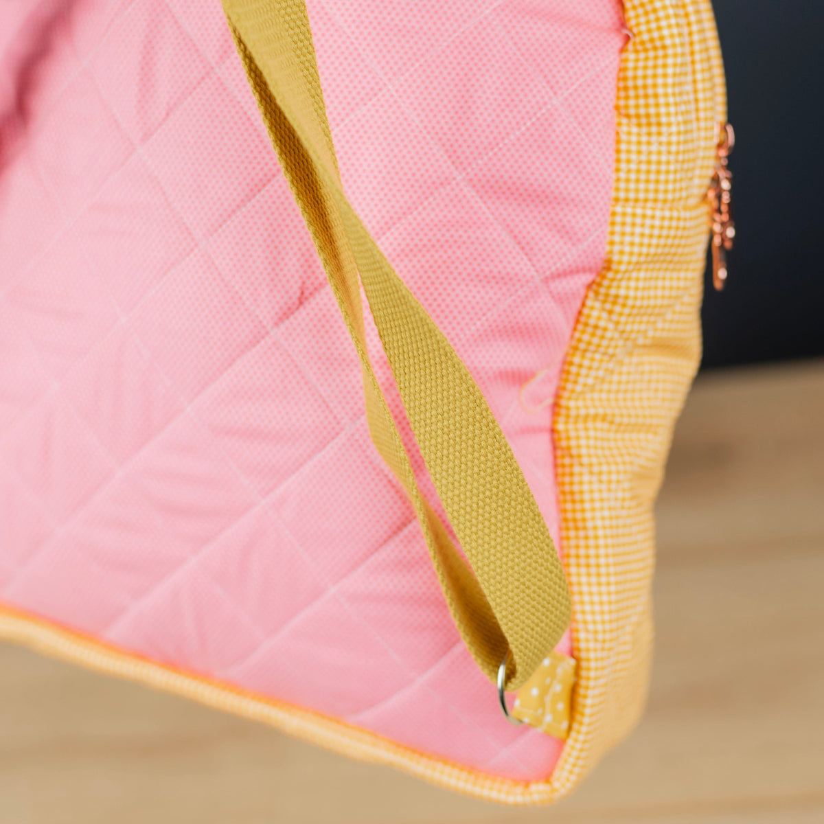 Close-up of a quilted pink bag with mustard yellow straps and a yellow-and-white gingham side panel, photographed against a dark background and light wood surface.