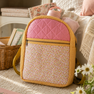A quilted pink and floral backpack with a yellow woven border, styled on a cream carpet next to a sofa with books and flowers.
