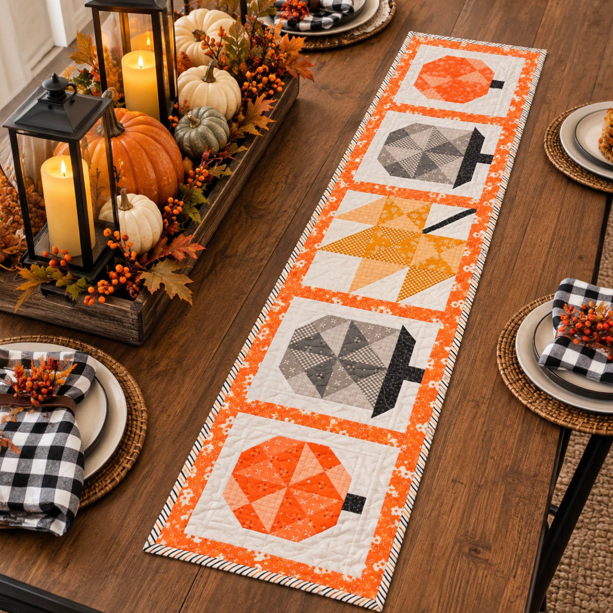 An autumn-themed table runner with four pumpkin and leaf quilted blocks in orange, yellow, gray, and white, styled on a wooden dining table with fall decorations and place settings.