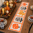 An autumn-themed table runner with four pumpkin and leaf quilted blocks in orange, yellow, gray, and white, styled on a wooden dining table with fall decorations and place settings.