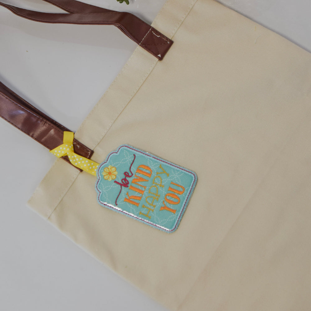 Beige tote bag with a colorful patch on a white surface with a plant and stack of books.