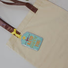 Beige tote bag with a colorful patch on a white surface with a plant and stack of books.