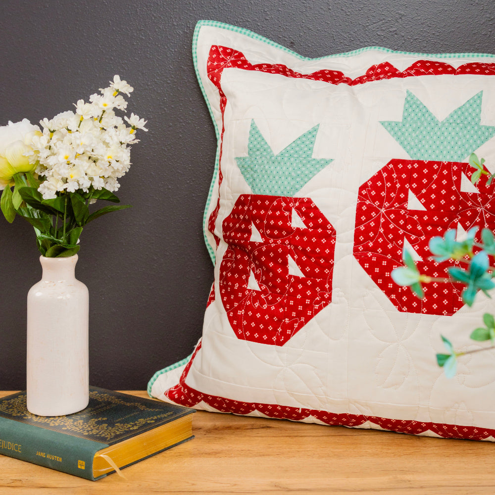 Decorative pillow with red and green patterns on a wooden surface next to a vase with flowers and a book.