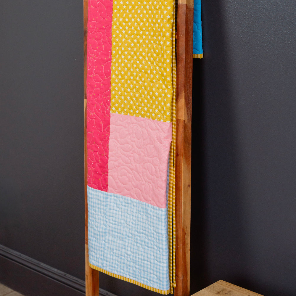 Colorful quilt with pink, yellow, and blue squares on a wooden ladder against a dark gray wall.
