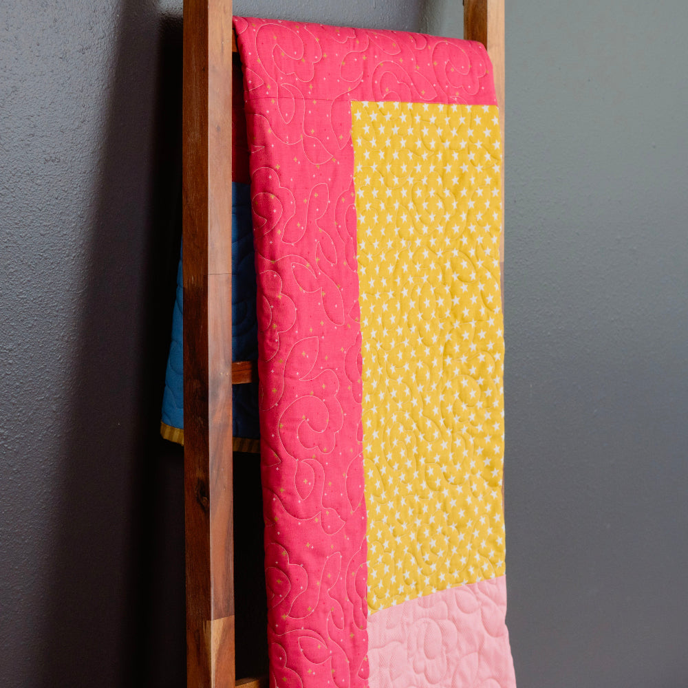 Colorful quilt with pink and yellow patterns draped over a wooden ladder against a gray wall.