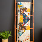 Colorful quilt displayed on a wooden ladder against a dark wall with a plant and small table in the foreground.