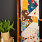 Colorful quilt on a wooden ladder against a black wall with a plant on a table.