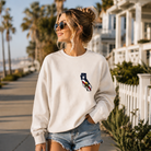 Woman wearing a white sweatshirt with a colorful California design on a beachside walkway.