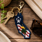 Keychain shaped like California with American flag design on a wooden surface with keys and sunglasses.