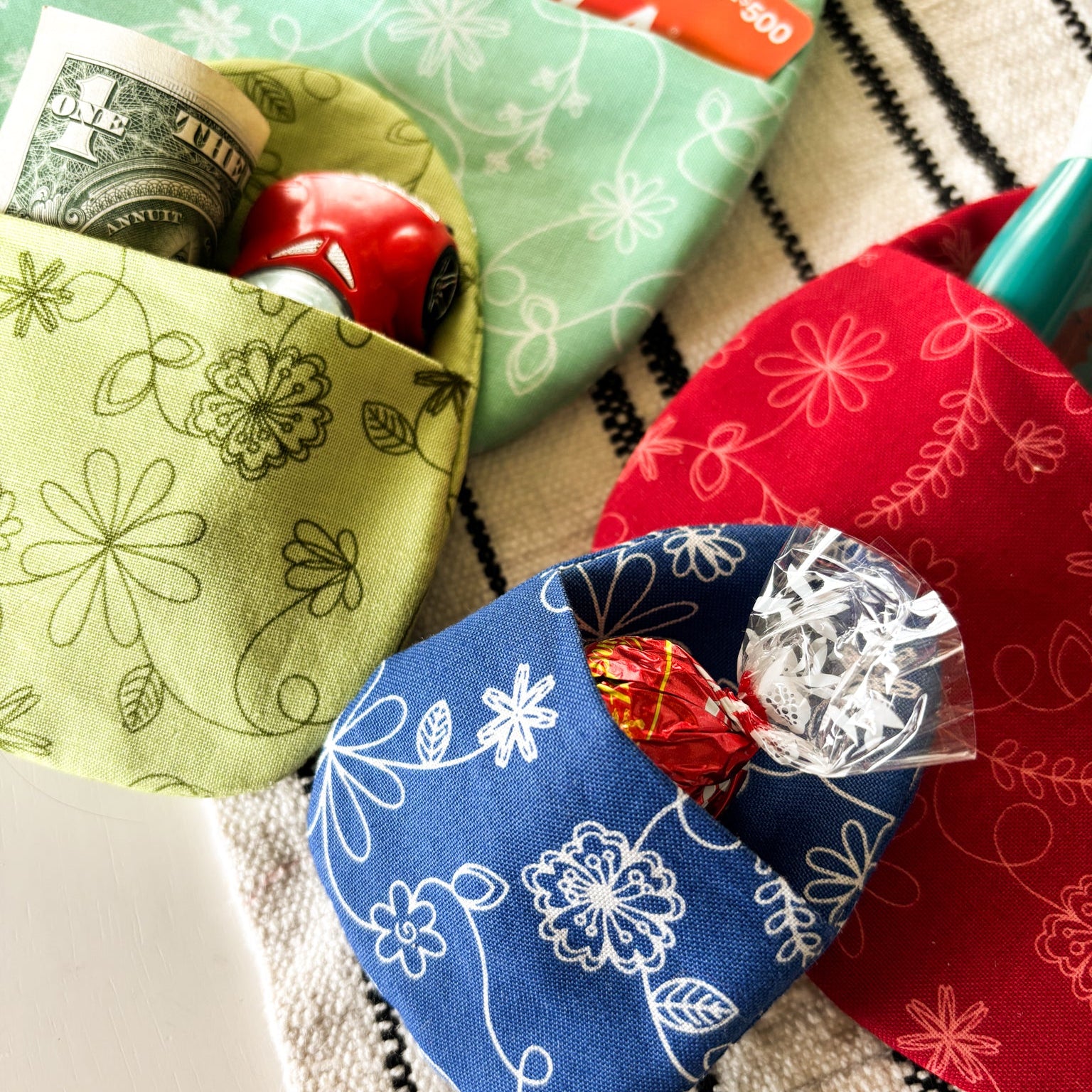 Three fabric pouches in green, blue, and red with candy and money on a striped background.
