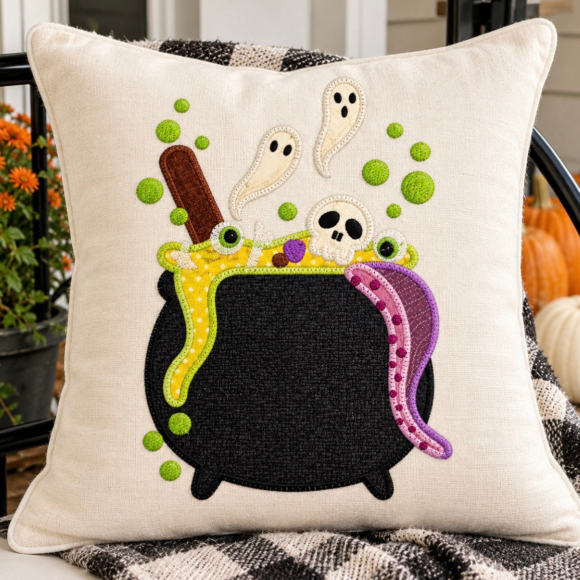 A square cream-colored linen throw pillow featuring a colorful embroidered Halloween cauldron design with ghosts, a skull, green potion bubbles, and a purple snake, styled on a porch chair with pumpkins and flowers in the background.