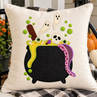 A square cream-colored linen throw pillow featuring a colorful embroidered Halloween cauldron design with ghosts, a skull, green potion bubbles, and a purple snake, styled on a porch chair with pumpkins and flowers in the background.