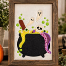 A framed Halloween-themed embroidered artwork displayed on a tabletop, surrounded by seasonal decor including a carved orange pumpkin, greenery, and a black-and-white buffalo plaid pumpkin. The artwork shows a black cauldron with green slime, white ghosts, a skull, colorful candies, and a purple snake on a cream fabric background inside a rustic wood frame.