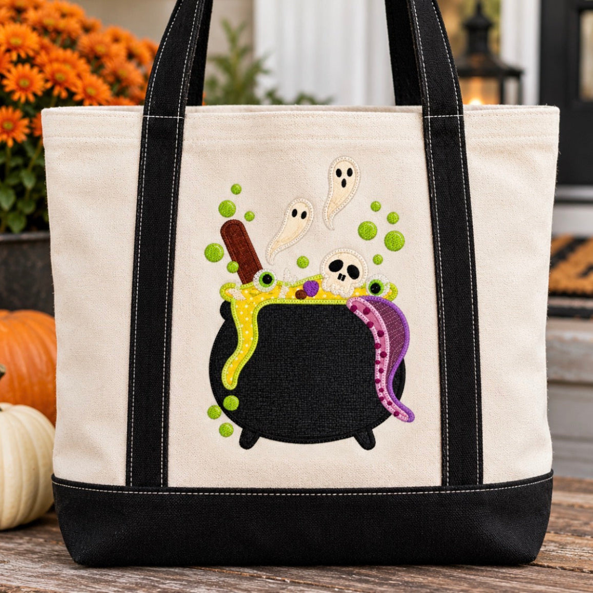 A canvas tote bag with black trim and handles, featuring a Halloween-themed applique design of a black cauldron, green bubbles, white ghosts, and a purple octopus on the front. The bag is styled on a wooden surface with pumpkins and orange flowers in the background.