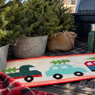 A rustic quilted table runner featuring cartoon-style Christmas trucks carrying green trees, styled in a pickup truck bed with potted Christmas trees and burlap.