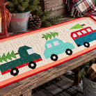 A quilted table runner featuring three Christmas-themed trucks in green, light blue, and red on a cream background with a coral border, styled on a rustic wooden table with a candle, pinecone, and plaid throw.