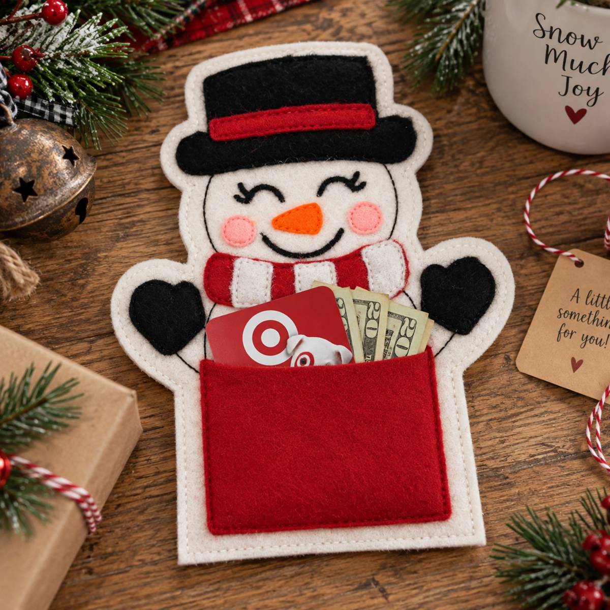 A festive felt snowman gift holder with a black top hat, red scarf, and red gift pocket, shown on a wooden table with holiday decorations, a wrapped gift, a mug, and a small tag.