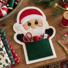 A festive felt Santa pocket organizer on a wooden table, filled with scissors, thread, and sewing accessories. The scene includes holiday decorations like pine branches, lights, patterned fabrics, and spools of thread.