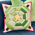 A square quilted throw pillow with a green and white gingham and floral patchwork design, featuring a central tan star and red button accents, resting on a blue couch with green pillows in the background.
