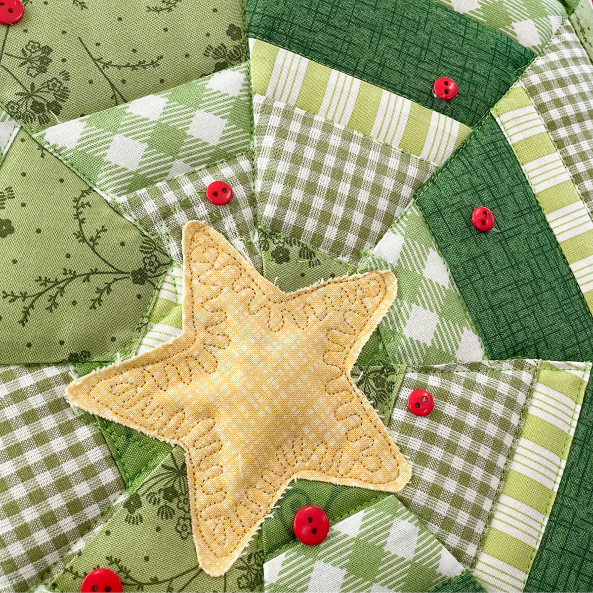 A close-up of a quilted fabric star patch in pale yellow with stitched leaf details, surrounded by green and white patterned quilted sections and small red buttons.