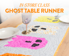 A quilted table runner displayed on a dining table, featuring three colorful ghost appliqués in pink, yellow, and white with black windowpane shapes. A wine glass and plates are styled on the table.