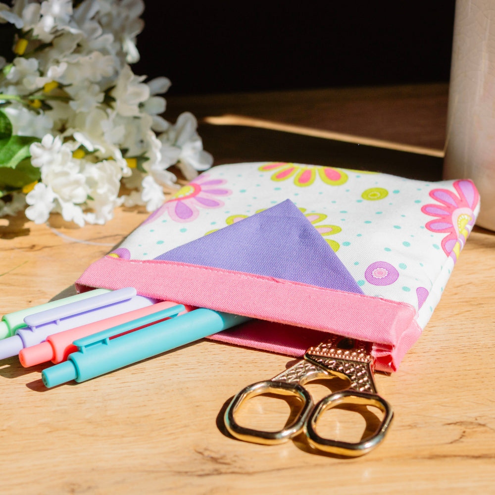 Colorful fabric pouch with markers and scissors on a wooden surface with flowers in the background
