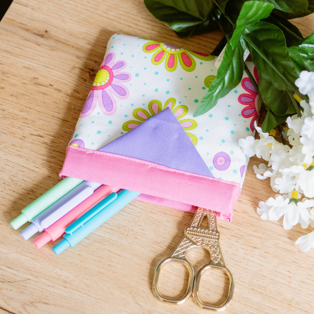Folded fabric with colorful flower patterns, straws, and scissors on a wooden surface with greenery and flowers.