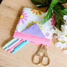 Folded fabric with colorful flower patterns, straws, and scissors on a wooden surface with greenery and flowers.