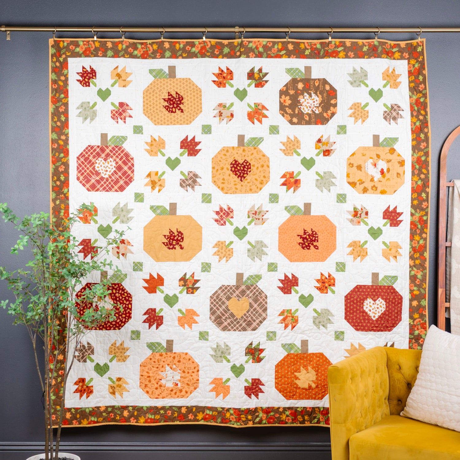 A large quilted wall hanging with an autumn pumpkin and leaf pattern, displayed on a dark gray wall in a styled room with a yellow armchair, a plant, and a wooden ladder.