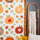 An autumn-themed quilted wall hanging or blanket featuring pumpkin motifs, fall leaves, and hearts in orange, red, yellow, green, and brown tones, styled in a room next to a wooden ladder shelf with a cream throw and a mustard yellow chair.