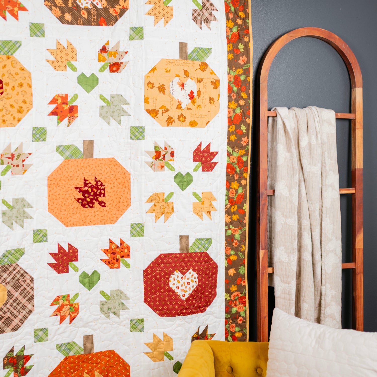 An autumn-themed quilted wall hanging or blanket featuring pumpkin motifs, fall leaves, and hearts in orange, red, yellow, green, and brown tones, styled in a room next to a wooden ladder shelf with a cream throw and a mustard yellow chair.