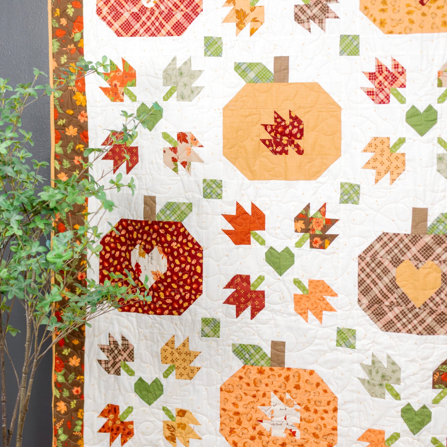A quilted fabric panel featuring autumn-themed patchwork designs including pumpkins, leaves, and hearts in orange, red, green, and brown on a white background, styled next to a small potted plant against a dark gray wall.