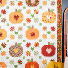 A handmade autumn-themed quilt hanging on a wall, featuring stitched pumpkins, leaves, and hearts in warm fall colors like orange, red, yellow, green, and brown on an off-white background.