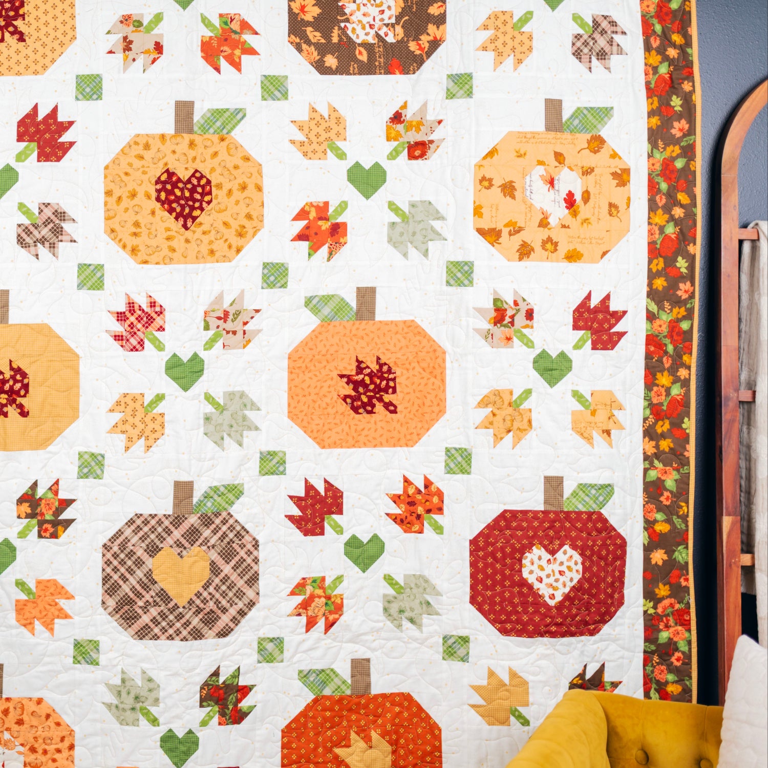 A handmade autumn-themed quilt hanging on a wall, featuring stitched pumpkins, leaves, and hearts in warm fall colors like orange, red, yellow, green, and brown on an off-white background.