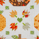 A close-up of a quilted fabric featuring an autumn-themed patchwork pattern with pumpkins, leaves, and hearts in orange, yellow, green, and brown tones on a white background.