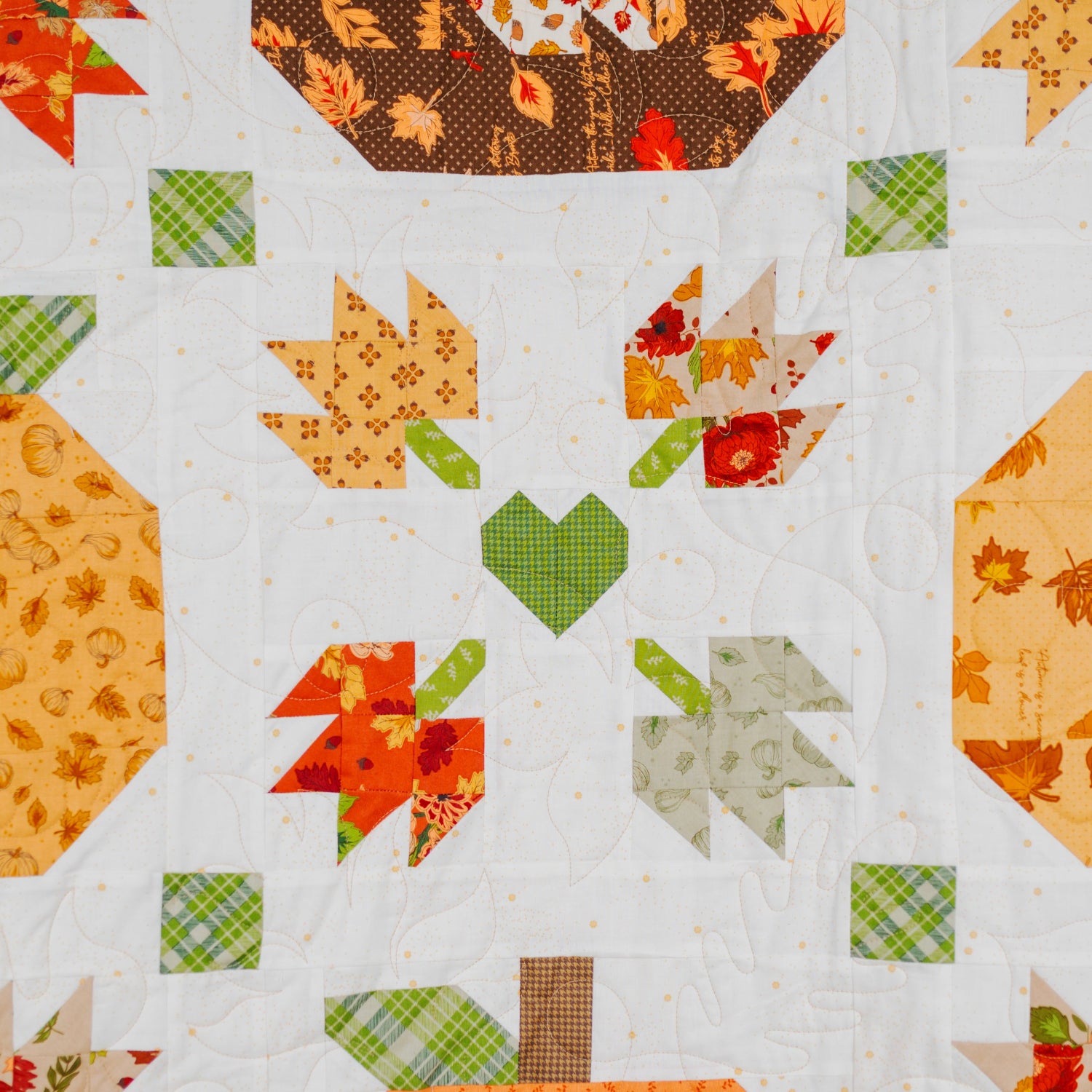 A close-up of a quilted fabric featuring an autumn-themed patchwork pattern with pumpkins, leaves, and hearts in orange, yellow, green, and brown tones on a white background.
