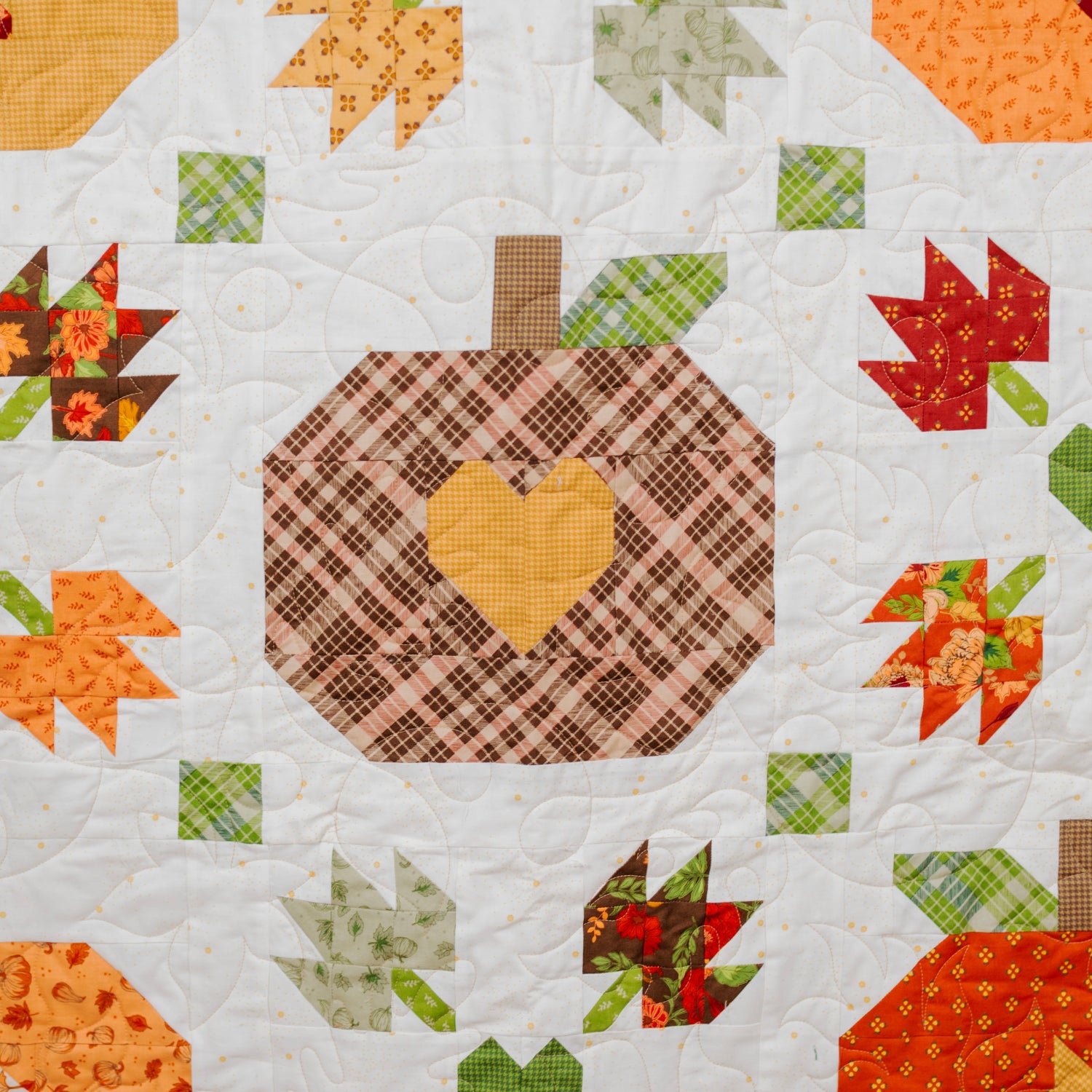 A close-up of a quilted fabric featuring an autumn harvest pattern with plaid pumpkins, colorful leaves, and green hearts on a white background.