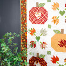 A close-up of a quilted fabric panel featuring autumn-themed pumpkin and leaf patterns in red, orange, green, and beige on a white background, with a dark gray wall and green foliage visible at the side.