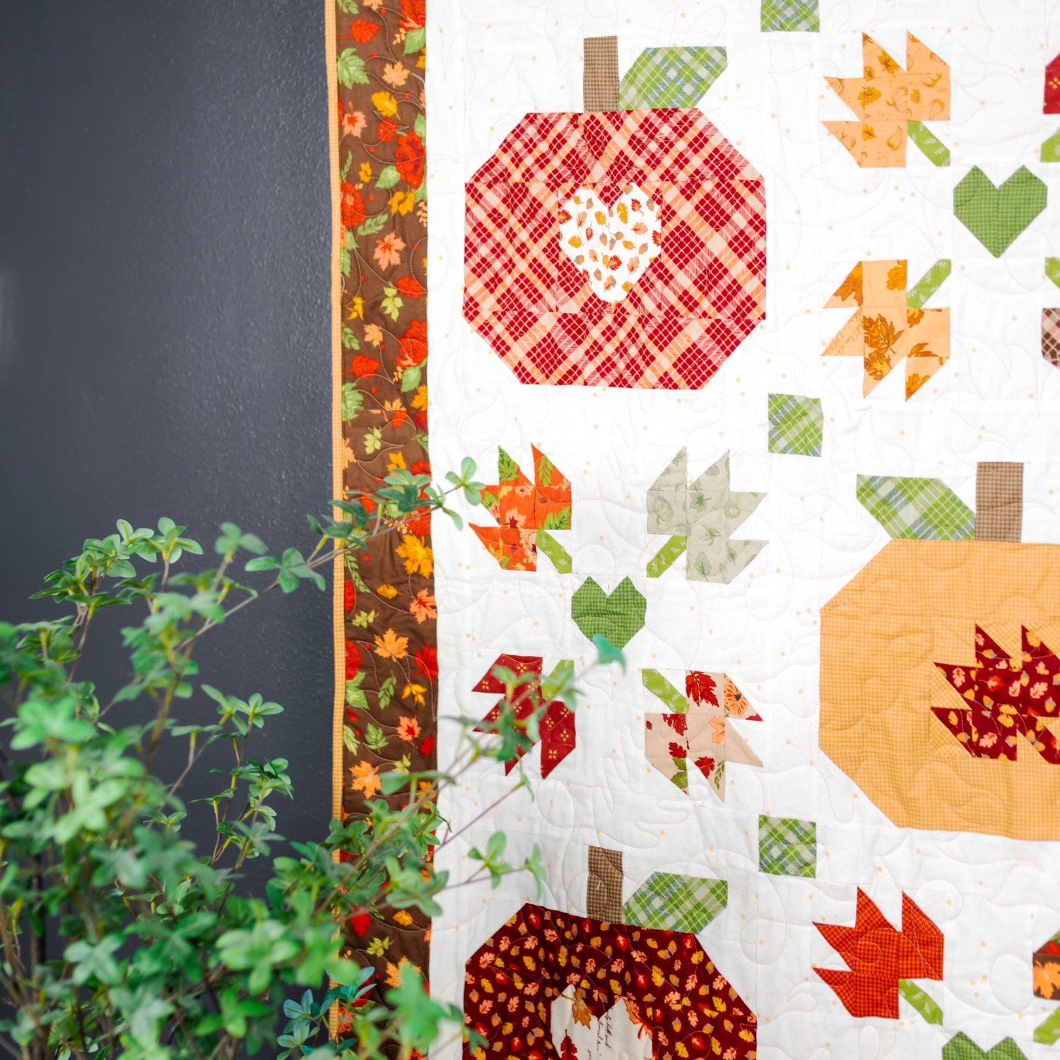 A close-up of a quilted fabric panel featuring autumn-themed pumpkin and leaf patterns in red, orange, green, and beige on a white background, with a dark gray wall and green foliage visible at the side.