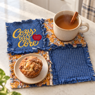 A set of four rustic blue fabric placemats with frayed edges and a floral patterned border, styled on a white marble kitchen counter with a mug of tea, a cinnamon stick, and a muffin.