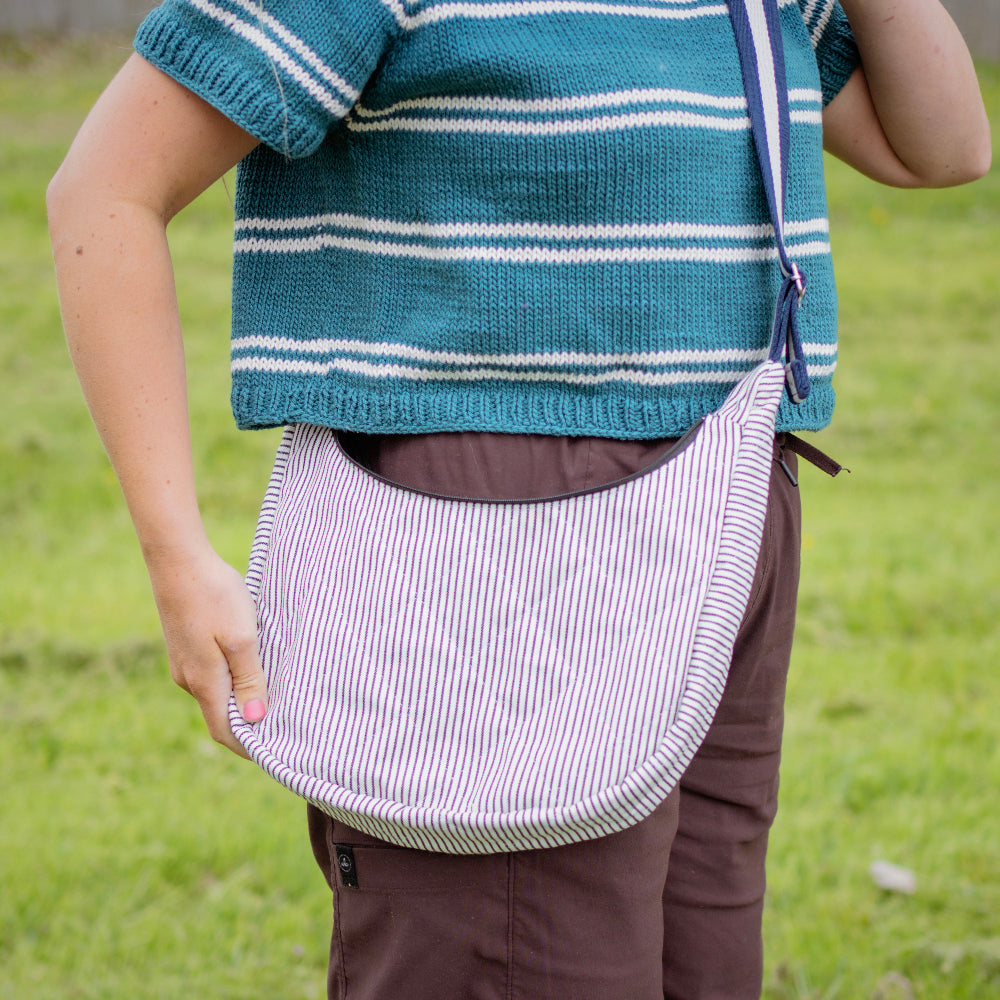 Person wearing a blue striped sweater and holding a white bag with black stripes in a grassy field.