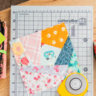 Fabric squares on a cutting mat with a rotary cutter, on a wooden surface.