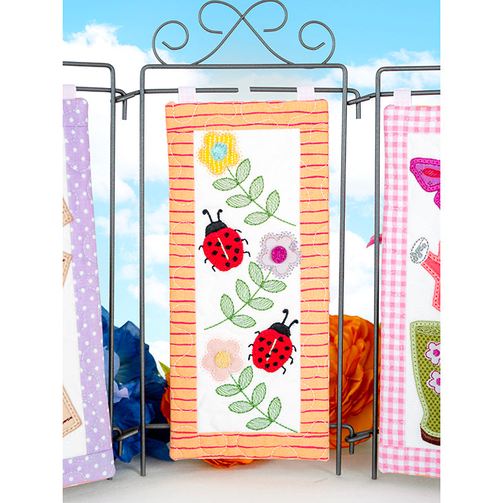 Embroidered fabric panels with ladybug and floral designs on a stand against a blue sky background.