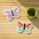 Two colorful butterfly coasters on a wooden surface with a green mug.